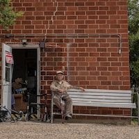 Boyd Stevens tenant sa canne, assis sur un banc à côté d'une porte portant un écriteau ''open'' d'un bâtiment délabré.