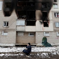 Une femme assise sur un trottoir porte les mains à son visage devant un immeuble brûlé.