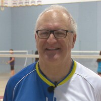 Il sourit à la caméra dans un gymnase où des jeunes jouent au badminton.