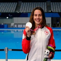 Aurélie Rivard pose avec sa médaille d'argent à la main sur le bord de la piscine. Elle sourit et porte une veste aux couleurs du Canada.