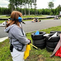 Une femme avec des écouteurs et un drapeau jaune dans les mains regarde une course de karting.