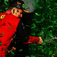 Un pilote dans une combinaison Ferrari secoue une bouteille de champagne, entouré de confettis verts. 