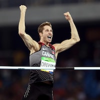 Un athlète lève les bras en triomphe devant la barre qu'il vient de franchir. 
