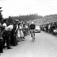 Image en noir et blanc d'un coureur cycliste qui monte une pente abrupte, entouré d'une foule compacte