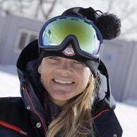 Une femme avec un manteau bleu marine et des lunettes de ski sur la tête sourit.