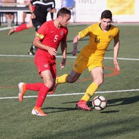 Un joueur de soccer en rouge tente de déjouer un adversaire en jaune qui le marque de près.