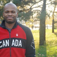 Un homme prend la pose pour la caméra devant des arbres et un rayon de soleil derrière. Il porte un manteau d'athlète du Canada.