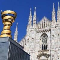 Un trophée est posée sur des caisses devant une cathédrale, sous un ciel bleu.