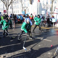Des syndiqués jouent au hockey-balle sur la Grande Allée en hiver avec, en arrière-plan, Bonhomme Carnaval et l’hôtel du Parlement.