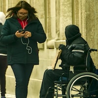 Une madame marche près d'une personne en fauteuil roulant en regardant son cellulaire. 