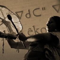 Une femme autochtone jouant du tambour; en arrière-plan, un texte écrit en cri.