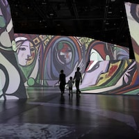 Un homme, une femme et un enfant se promènent dans l'exposition, entourés d'œuvres de Picasso projetées sur les murs.