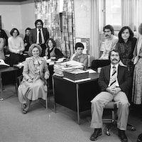 Des membres du personnel de la station C BON, douze hommes et femmes, posent dans les locaux, certains devant leur bureau, d'autres derrière. Photo en noir et blanc.