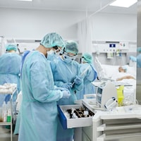 À l'avant-plan, deux professionnels de la santé manipulent des fournitures médicales. Derrière eux, d'autres s'occupent d'un patient dans un lit d'hôpital. Tous portent des vêtements protecteurs. 