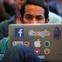 Un homme est derrière son ordinateur, assis au sein d'une foule; des étiquettes de Facebook et de Google sont collées sur le couvercle de son portable.