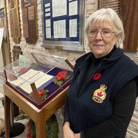 Gisèle Pharand se tient devant un mur rempli de plaques, et il y a un cartable dans une caisse vitrée à ses côtés.