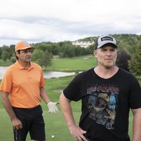 Deux hommes sur un terrain de golf, l'un habillé en orange et l'autre avec un gilet noir et une casquette grise.