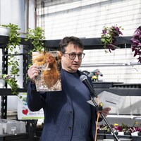 Un homme Eddy Métivier,  qui parle au micro, devant des étagères de plantes. Il tient un guide sur l'élevage des poules dans sa main.