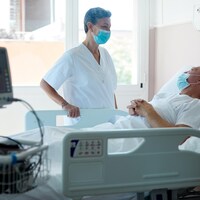 Une infirmière masquée parle à un patient alité dans une chambre d'hôpital. 