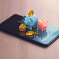 Des petits cochons tirelires d'où sortent des pièces de monnaie posés sur l'écran d'un cellulaire qui affiche les résultats de marché financier. 