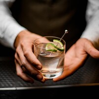 Gros plan sur les mains d'un homme qui tend un cocktail servi dans un verre et décoré d'une tranche de lime.
