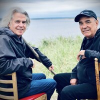 Deux hommes vêtus de vestes sombres sont assis sur deux chaises sur une plage. Derrière eux, on peut voir la mer.