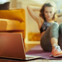 Une femme fait des abdos sur un tapis de yoga dans son salon en regardant un écran d'ordinateur.
