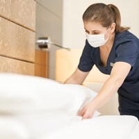Une femme de ménage portant un masque fait un lit dans une chambre d'hôtel.