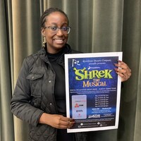 Une jeune femme souriante aux cheveux tressés qui porte des lunettes, tient une affiche sur laquelle est inscrite Shrek The Musical.