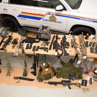 Sur une table devant un véhicule de la GRC des d’armes d’épaules, des gilets pare-balles, des armes de poing, une grande quantité de munitions et une machette.
