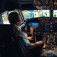 Plan large de la cabine de pilotage avec tous les cadrans, écrans et boutons. Un pilote est assis dans son siège et consulte les écrans.