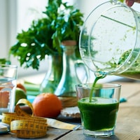 Un pichet qui verse un jus vert dans un verre sur une table entouré d'autres aliments et d'un ruban à mesurer.