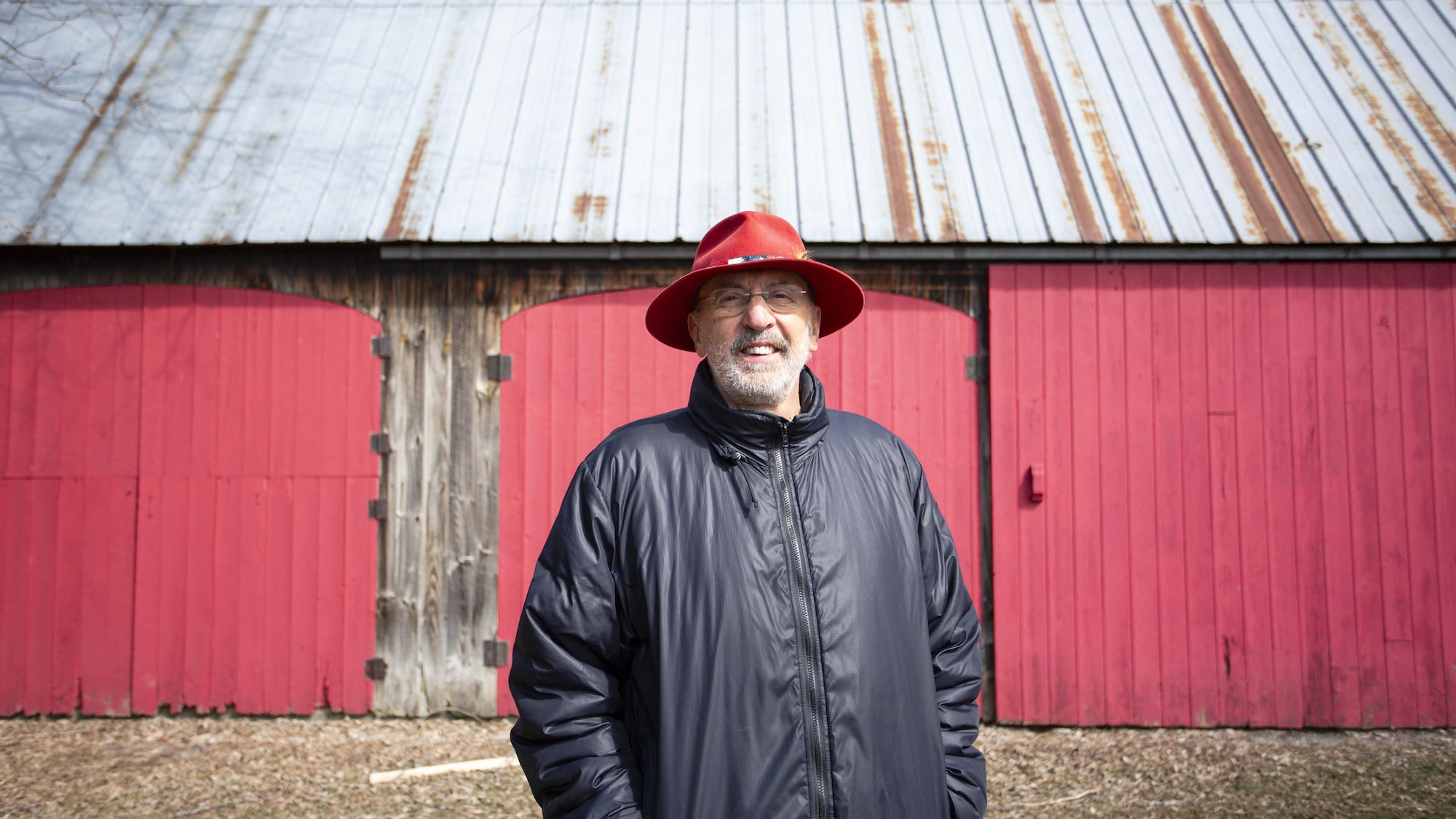 Gaston Lepage se tient devant une grange en bois, dont les portes sont peintes en rouge. Il sourit et porte un chapeau à large rebords rouge.