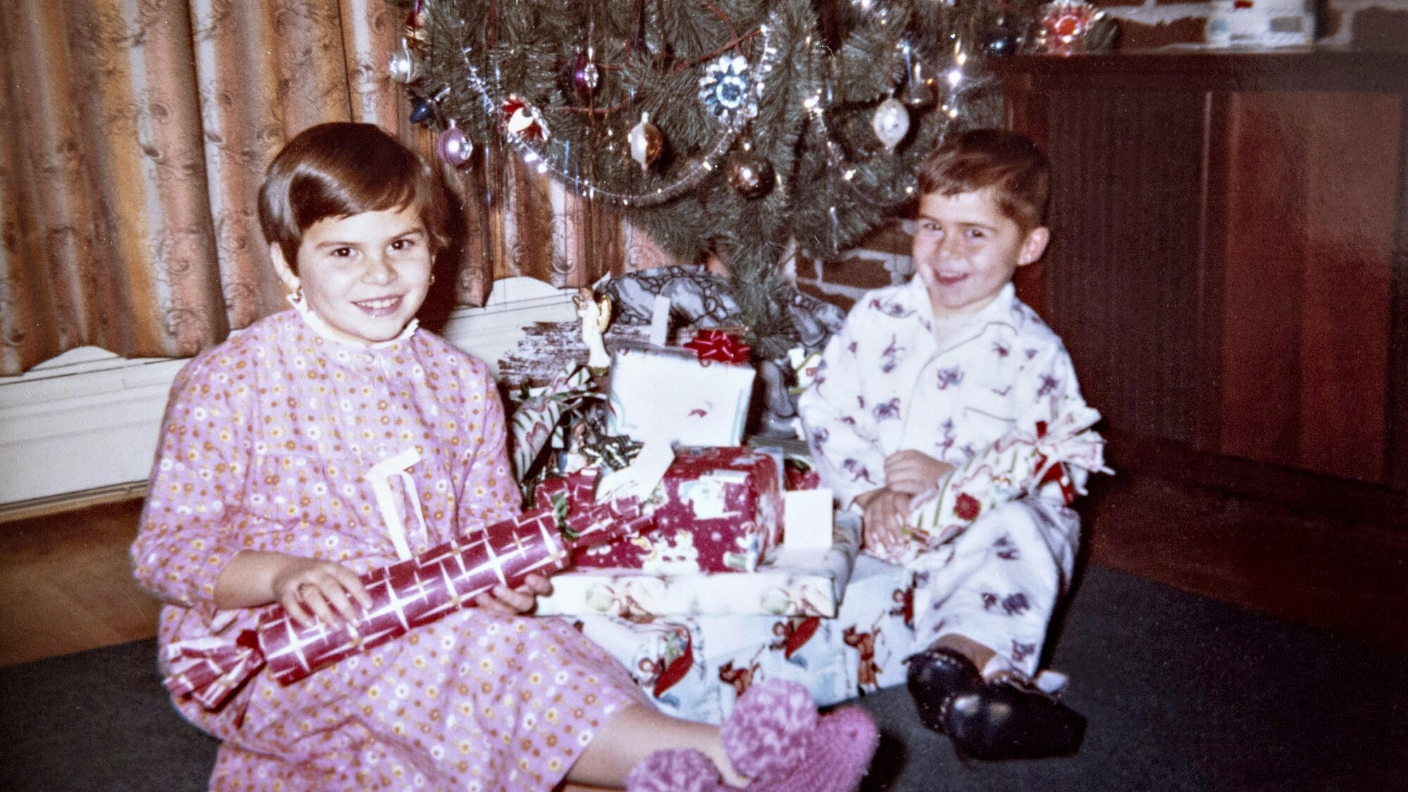 Une jeune fille et son frère sont assis sur le plancher en pyjama, devant un sapin de Noël.