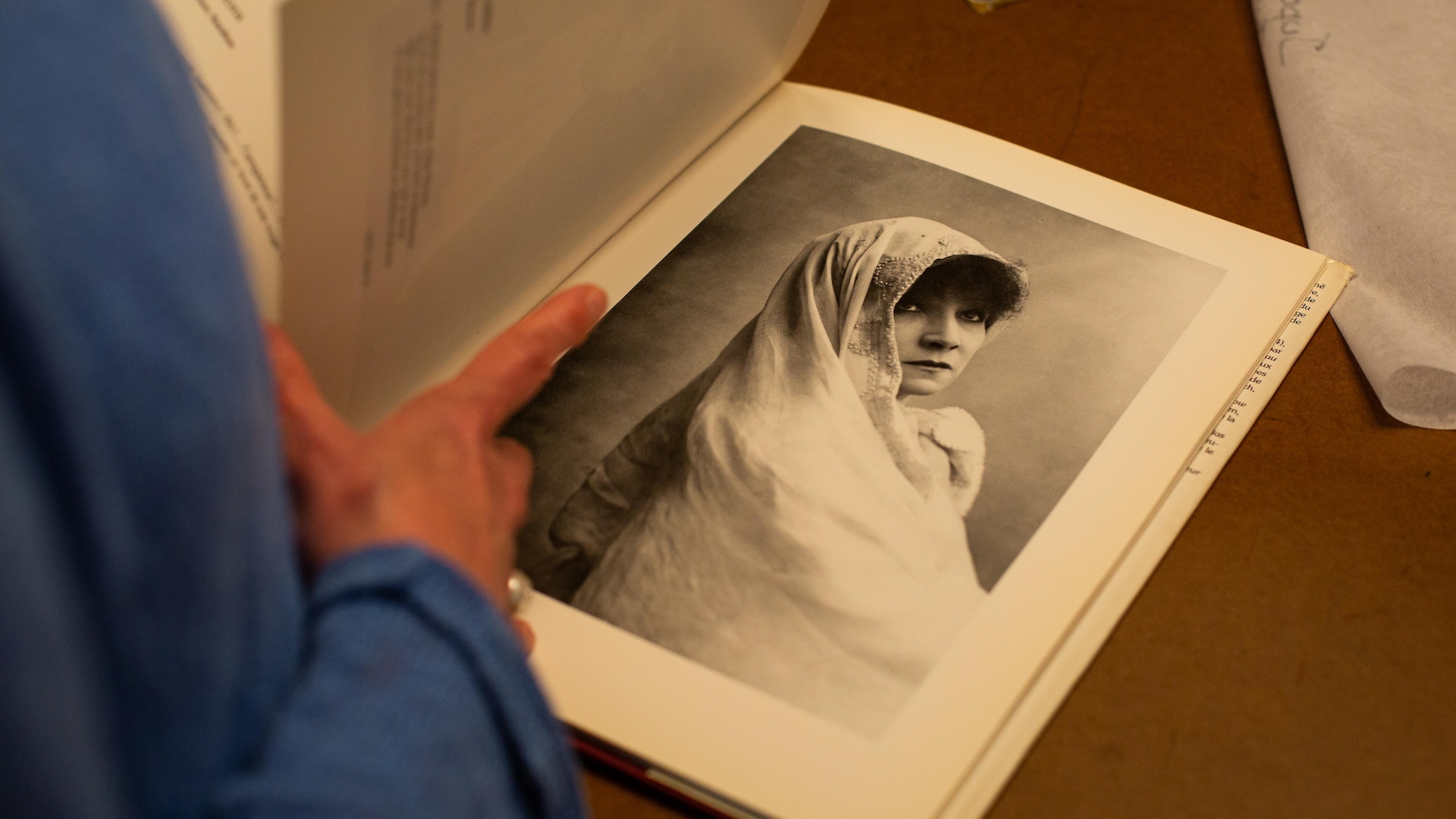 Marie-Chantal Perron feuillette un livre illustré. Sur la photo, la page montre une photo en noir et blanc de Sarah Bernhardt avec un voile blanc sur la tête.
