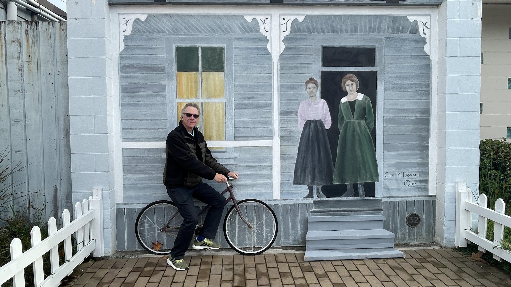 Un homme sur bicyclette pose devant une murale sur laquelle deux femmes dont debout sur un balcon.