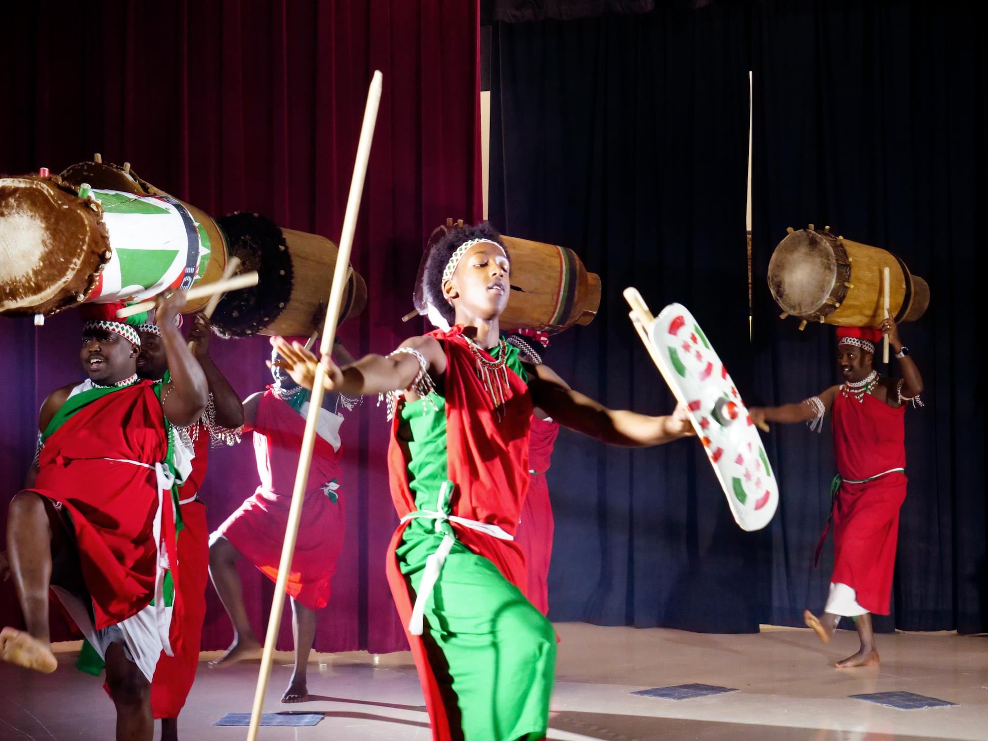 Sur scène, un jeune garçon en tenue traditionnelle, vu de dos, tient des baguettes en bois face à un grand tambour décoré du drapeau burundais. Derrière lui, plusieurs hommes adultes en toges rouges et vertes sont alignés, debout derrière leurs propres tambours en bois.