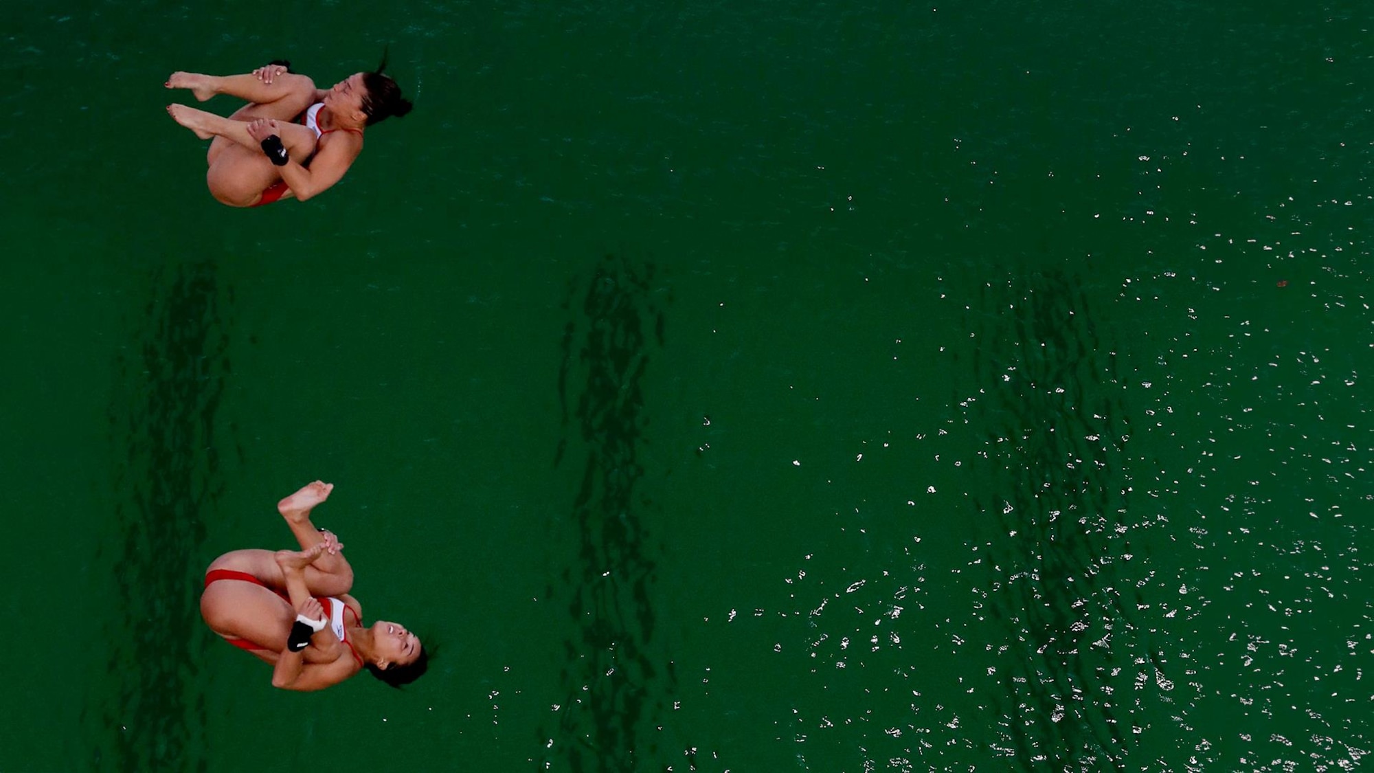 Deux plongeuses font des figures synchronisées devant un plan d'eau verte