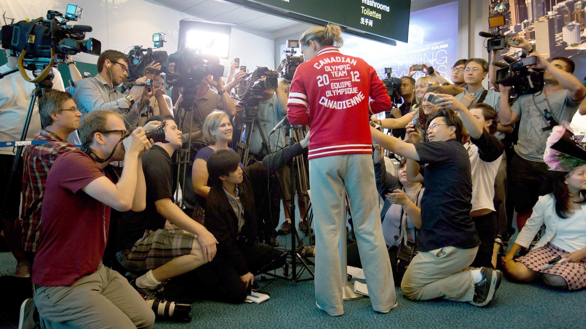 Une femme avec une veste rouge, de dos, répond à des questions dans une mêlée de presse avec de nombreux journalistes