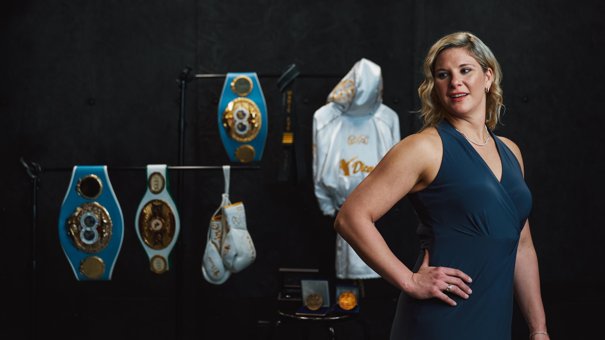 Elle prend la airs en robe de soirée devant des objets relatant sa carrière en boxe
