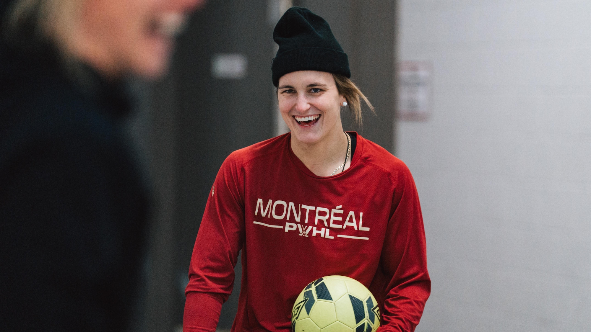 Une femme avec un chandail rouge et une tuque noire sourit avec un ballon de soccer dans les mains. 