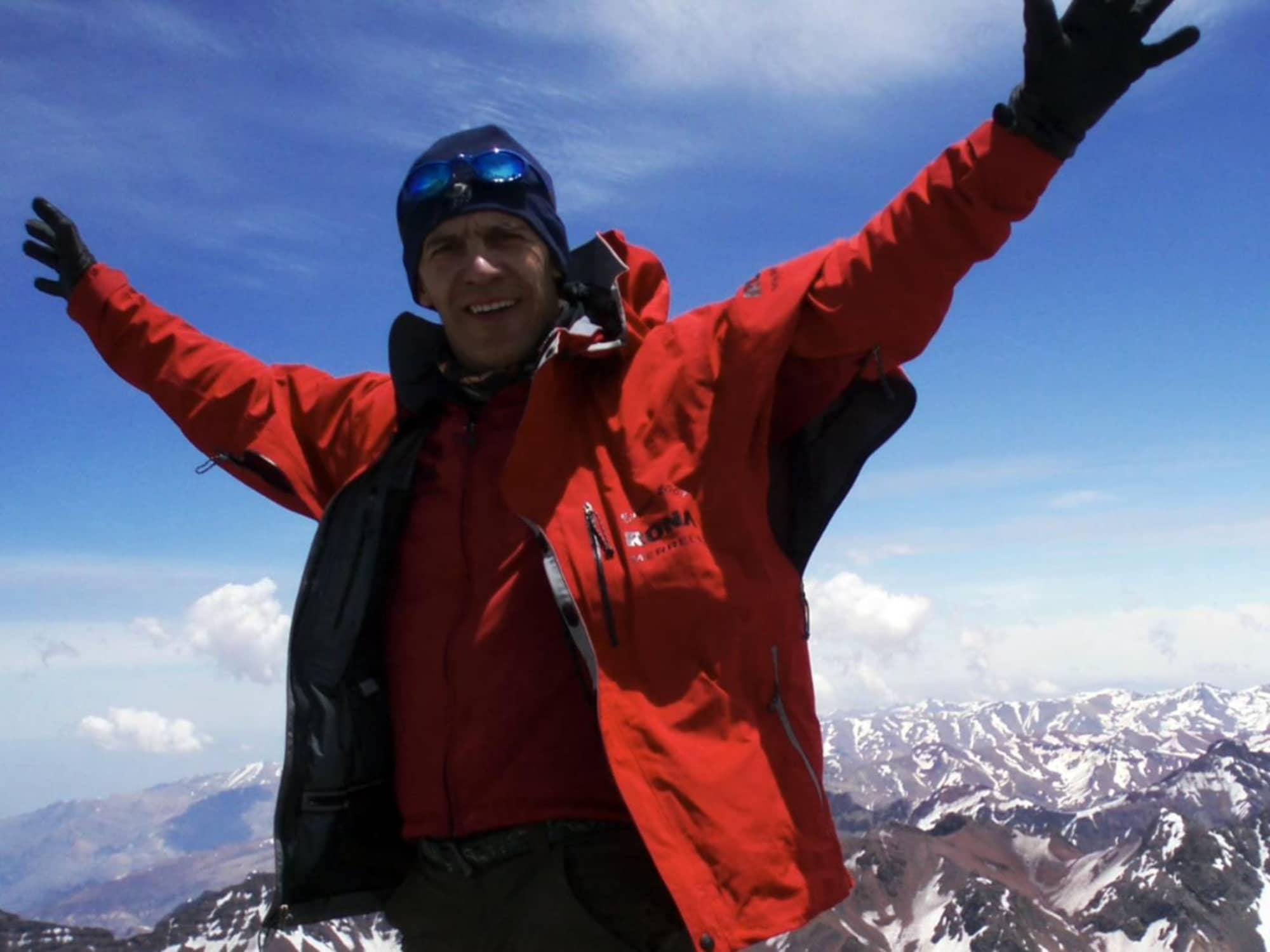 Un homme au sommet d'une montagne lève les bras en souriant.