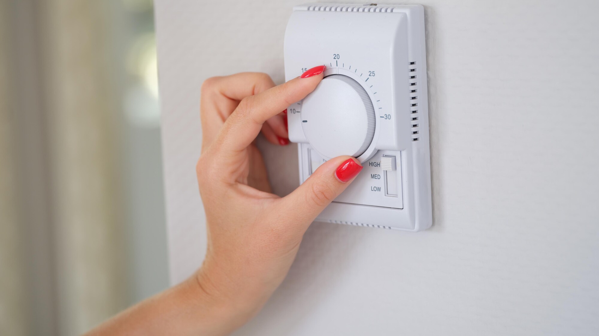 Plan rapproché de la main d'une femme aux ongles vernis qui tourne la molette d'un thermostat.