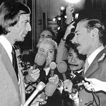 Cesar Menotti est devant Jorge Videla et plusieurs personnes qui tiennent des microphones.