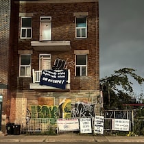 Un triplex abandonné. Sur le balcon, une banderole avec l'inscription « Logements vides, logements chers, on occupe! ». Une clôture devant l'immeuble pour empêcher d'accéder au terrain.