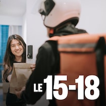 Dans l'embrasure d'une porte, une femme sourit à la personne qui vient lui livrer sa nourriture tout en prenant le sac en papier brun qui lui est tendu.