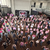 Une vue en plongée montre une foule nombreuse participant à une danse en ligne dans une grande salle. La majorité des danseurs portent des hauts roses. Une scène avec de l'équipement de sonorisation est installée au fond de la pièce.
