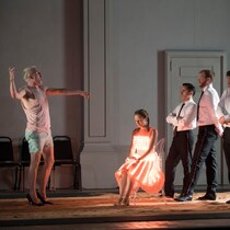 Quatre hommes en costume regarde un homme portant un débardeur, un short et des talons hauts en train de chanter, pendant qu'une femme est assise sur une chaise..