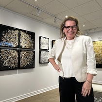 L'artiste porte des lunettes noires, sourit, et se tient à côté de ses toiles abstraites.