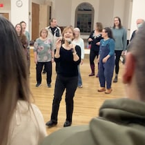 Une femme tient un micro et se trouve au centre d'un cercle de chanteurs amateurs.